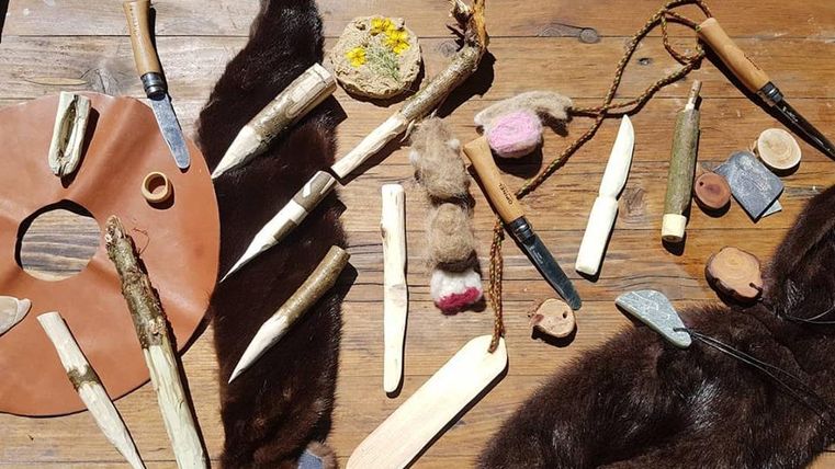 Eine Vielzahl von handgefertigten Werkzeugen und Materialien liegt auf einem Tisch. Darunter sind Holzstücke, Steinwerkzeuge und ein Lederstück zu sehen.