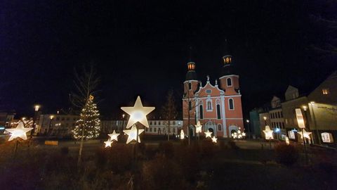 A festive city view at night with a decorated Christmas tree and glowing star figures. In the background stands a beautiful church in vibrant colors.