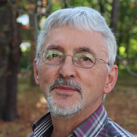 Ein älterer Mann mit grauen Haaren und Brille lächelt in die Kamera. Der Hintergrund ist unscharf und zeigt Bäume und herbstliche Farben.