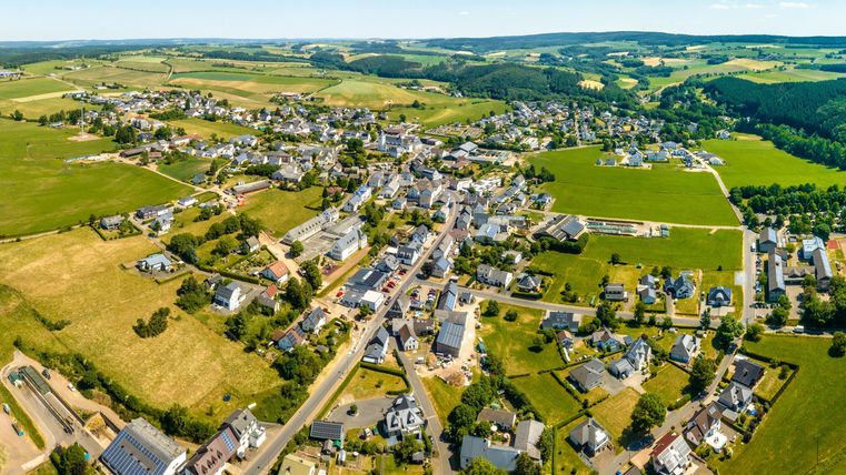 Une vue aérienne d'un petit village entouré de champs verts et de collines. Les maisons sont dispersées et entre elles, des routes et des espaces naturels sont visibles.