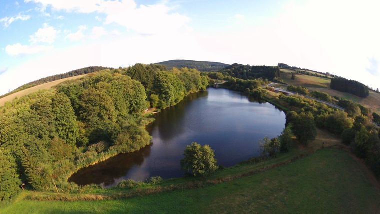Ein ruhiger Teich, umgeben von grünen Bäumen und sanften Hügeln. Der Himmel ist teilweise bewölkt, was der Szenerie eine friedliche Atmosphäre verleiht.
