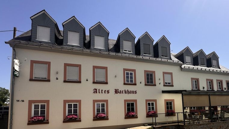 Ein historisches Gebäude mit dem Namen "Altes Bachhaus". Die Fassade ist hell, mit bunten Blumenkästen und einem klaren blauen Himmel im Hintergrund.