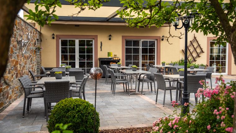 Eine gemütliche Terrasse mit Sitzgelegenheiten und Tischen. Umgeben von grünen Pflanzen und bunten Blumen.