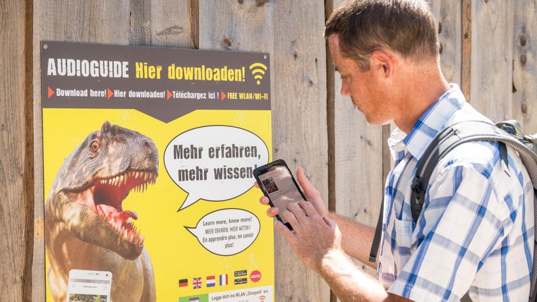 Un homme scanne un code QR sur une affiche avec son smartphone. L'affiche contient des informations sur des audioguides et montre une image d'un dinosaure.