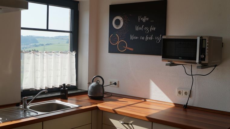 Eine helle Küche mit modernen Möbeln und einem Fenster, das Tageslicht hereinscheinen lässt. Im Hintergrund sind ein Mikrowelle und eine kreative Wanddekoration zu sehen.