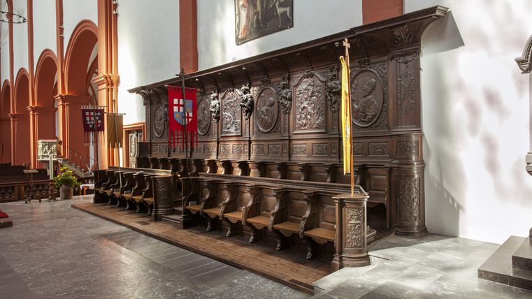 Eine detaillierte Holzbank in einer Kirche mit geschnitzten Ornamenten. Im Hintergrund sind weitere architektonische Elemente der Kirche sichtbar.