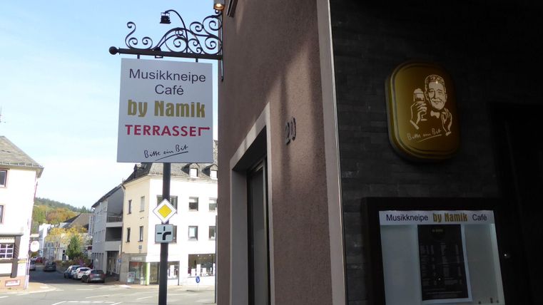 Ein Schild für eine Musikkneipe und Café mit dem Namen "by Namik" ist an der Wand angebracht. Es ist eine ruhige Straße mit mehreren Gebäuden im Hintergrund.