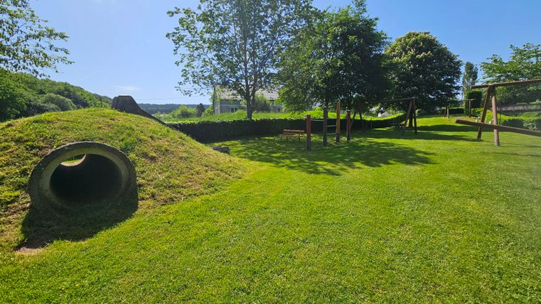 Ein großer, grüner Spielplatz mit einer Hügelstruktur und einem Tunnel. Im Hintergrund sind Bäume und Spielgeräte zu sehen.