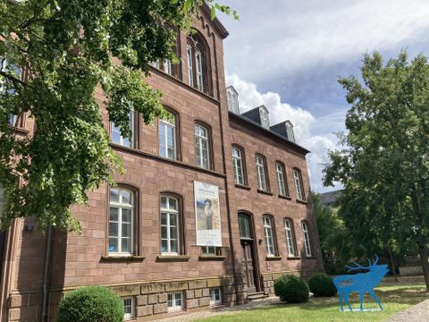 Ein historisches Gebäude aus rotem Stein mit großen Fenstern. Umgeben von Bäumen und grünem Rasen unter einem blauen Himmel.