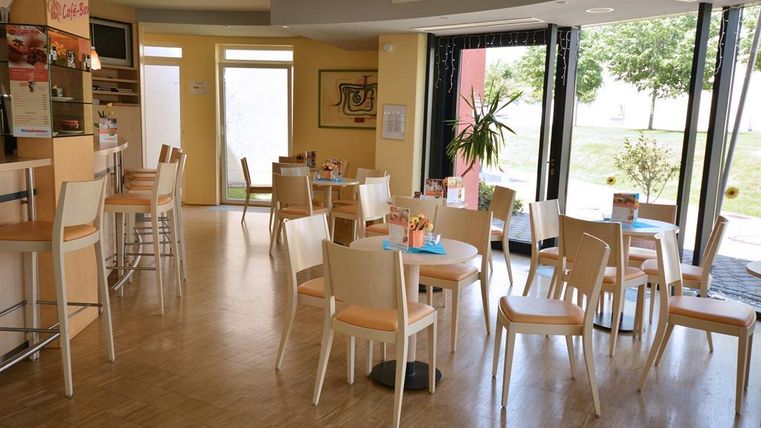 Ein helles Café mit mehreren Tischen und Stühlen. Große Fenster bieten einen Blick auf das Grün draußen.