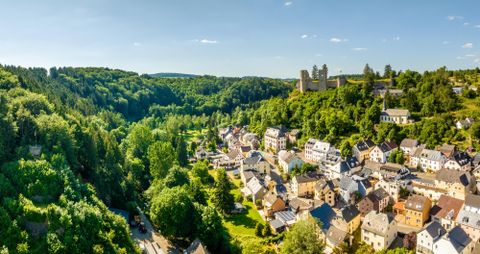 A picturesque landscape with a small village surrounded by lush greenery. In the background, a castle ruin is visible on a hillside.