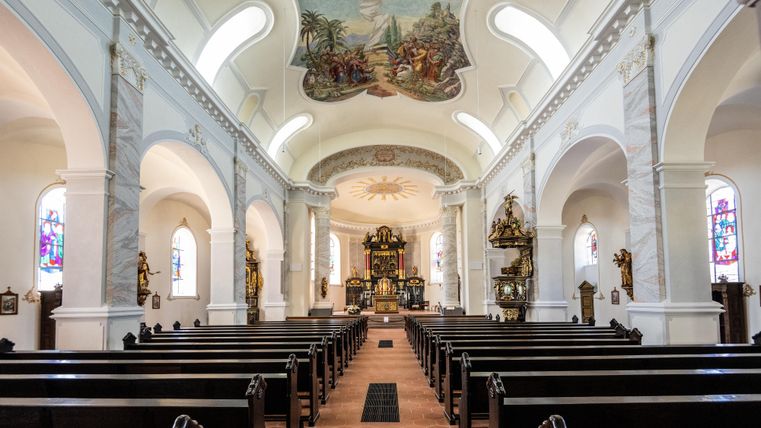 A bright church with high ceilings and colorful windows. The benches are neatly lined up and an impressive altar arrangement is visible.