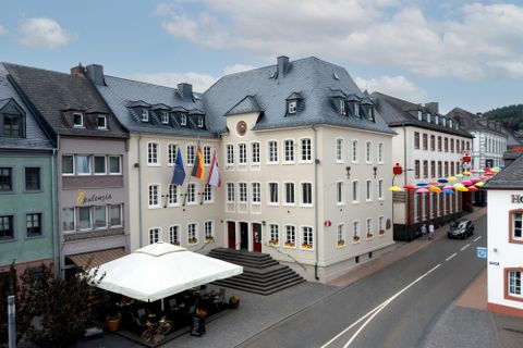Ein historisches Gebäude mit mehreren Fahnen vor der Fassade. Auf der Straße sind bunte Wimpel und ein Restaurant mit Sonnenschirm zu sehen.
