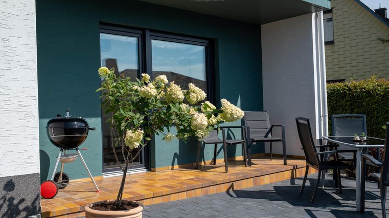 Eine gemütliche Terrasse mit modernen Möbeln und einem Grill. Vor der Terrasse blühen weiße Pflanzen im Topf.