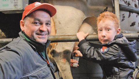 Ein Bauer und ein Junge stehen neben einer Kuh in einem Stall. Sie lächeln in die Kamera und zeigen eine enge Verbindung zur Landwirtschaft.