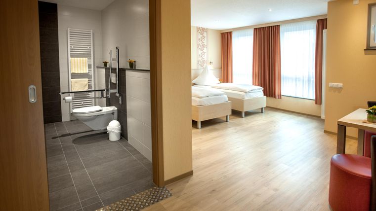 Ein modernes Hotelzimmer mit zwei Einzelbetten und großen Fenstern. Im Hintergrund ist ein Badezimmer mit modernen sanitären Einrichtungen sichtbar.