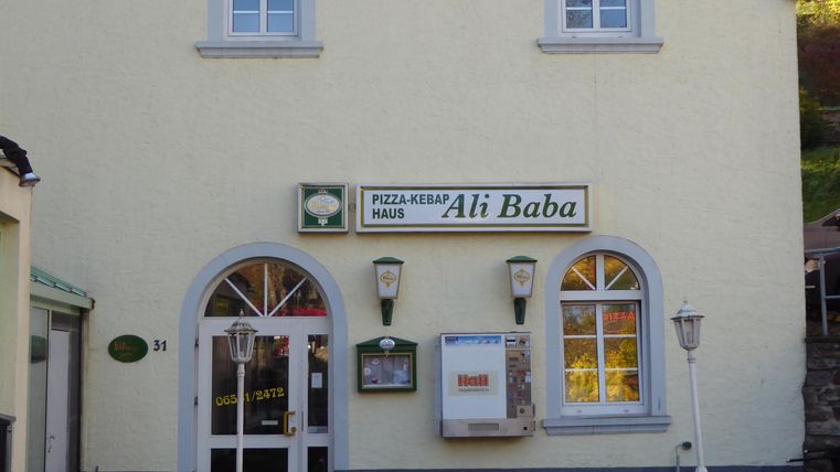 Ein Restaurant mit der Bezeichnung "Ali Baba" ist an einer hellen Gebäude-Fassade zu sehen. Die Fenster sind oval und die Eingangstür lädt zum Betreten ein.