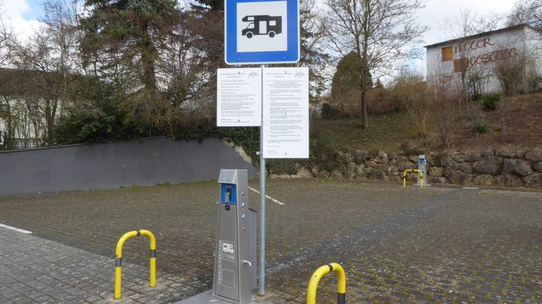 Ein Parkplatz für Wohnmobile mit einer Informationsbeschilderung. Im Vordergrund steht ein Zahlungsterminal neben gelben Pollern.