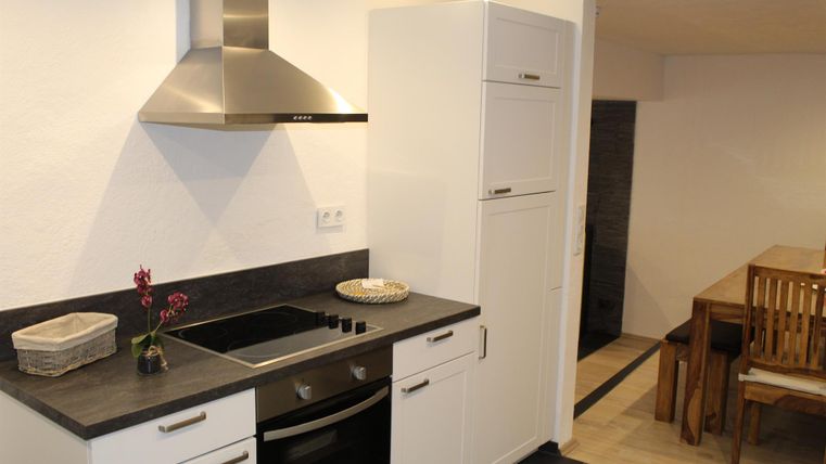 Eine moderne Küche mit einer Edelstahlabzugshaube, einem Herd und weißen Schränken. Im Hintergrund befindet sich ein Holz-Essbereich mit Tisch und Stühlen.