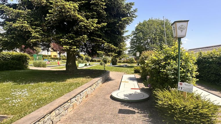 Un terrain de mini-golf bien entretenu avec des pelouses vertes et des arbres. Le temps est ensoleillé et bleu, idéal pour une visite de loisirs.