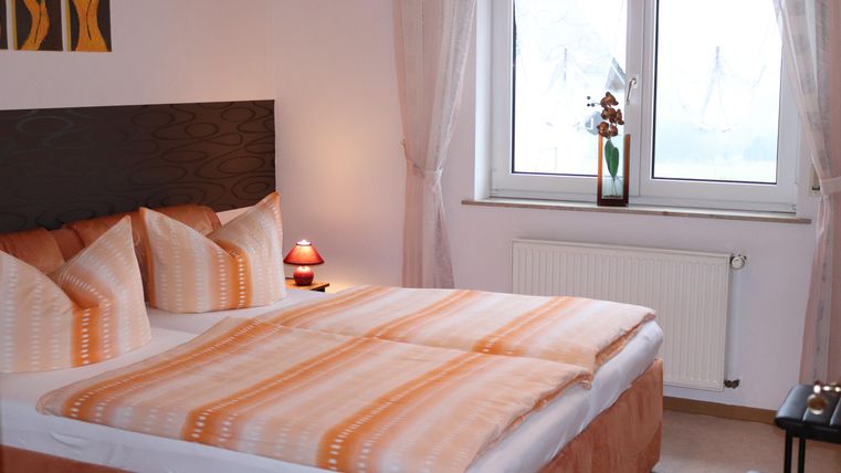 Ein stilvolles Schlafzimmer mit einem großen Doppelbett und orangefarbenen Bettdecken. Helle Fenster sorgen für eine freundliche Atmosphäre und eine angenehme Einrichtung.