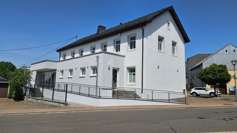 Une maison moderne de deux étages avec une façade blanche. Elle est située le long d'une rue et dispose d'une entrée accessible.