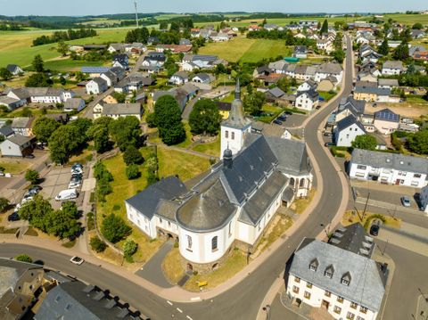 Eine Luftaufnahme eines kleinen Dorfes mit einer Kirche im Mittelpunkt. Die Umgebung ist von grünen Feldern und Wohnhäusern umgeben.
