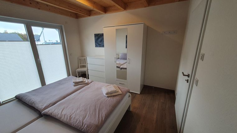 Ein modernes Schlafzimmer mit zwei Betten, einem Kleiderschrank und einem Stuhl. Große Fenster sorgen für viel natürliches Licht.