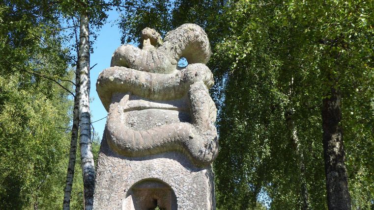 An impressive stone sculpture depicting a snake encircles a central block. Surrounded by green trees and a tranquil setting.