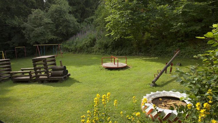 Ein Spielplatz mit verschiedenen Spielgeräten in einem grünen Park. Die Umgebung ist von Bäumen und Blumen umgeben.