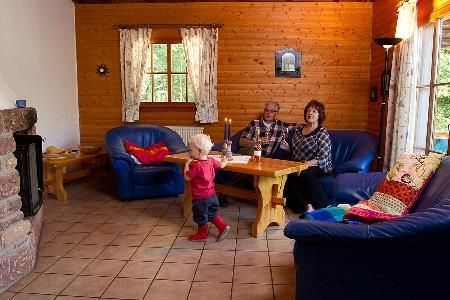 Ein gemütliches Wohnzimmer mit Holzverkleidung und zwei blauen Sofas. Ein Kind spielt, während ein Paar am Tisch sitzt und sich unterhält.