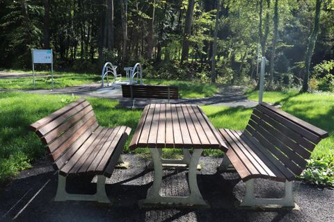 Ein gepflasterter Sitzbereich mit Holzbänken und einem Tisch im Wald. Im Hintergrund sind Grünflächen und Spielgeräte zu sehen.
