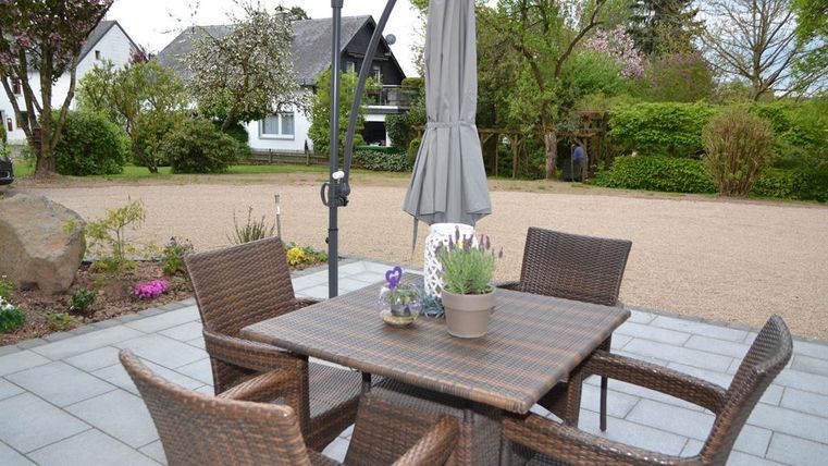 Ein gemütlicher Außenbereich mit einem Tisch und Stühlen aus Rattan. Im Hintergrund sind Bäume und ein Wohnhaus zu sehen.