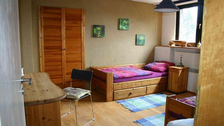 Ein einfaches Zimmer mit Holzmöbeln und einem Einzelbett. Die Wände sind beige, und es gibt einige grüne Bilder und einen Teppich in bunten Farben.