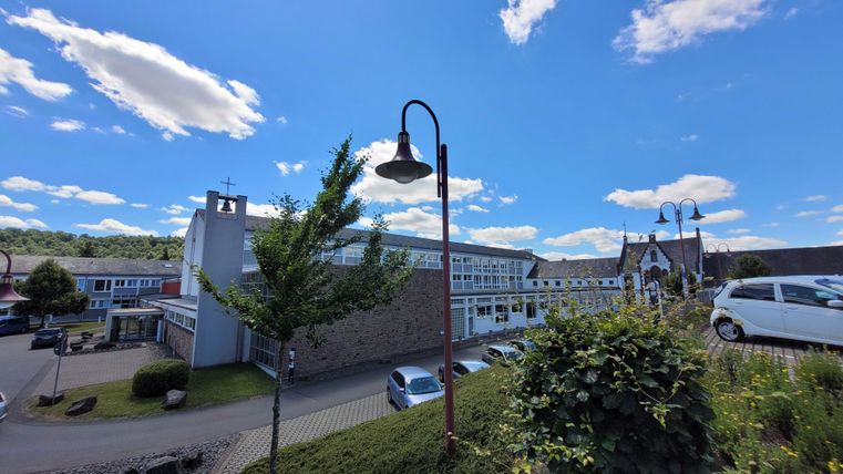 Un endroit paisible, sous un ciel bleu limpide parsemé de quelques nuages. Au premier plan, on aperçoit des arbres et un lampadaire, tandis que des bâtiments se dessinent à l'arrière-plan.