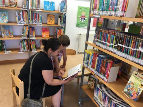 Eine Frau und ein Kind sitzen in einer Bibliothek und schauen gemeinsam in ein Buch. Im Hintergrund stehen Regale voller Bücher.