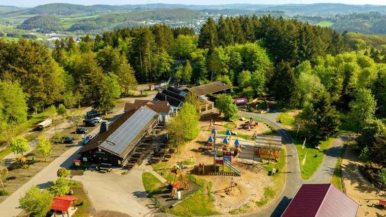 Une vue aérée d'une aire de jeux entourée d'arbres et de cabanes. L'environnement accueillant invite les familles à se détendre et à jouer.