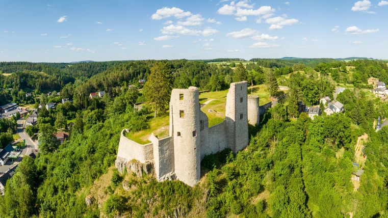 An old castle ruin on a hill, surrounded by green forests. In the background, small houses and a clear sky can be seen.