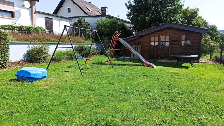 Ein Spielplatz mit einer Schaukel und einer Rutsche steht auf einer grünen Wiese. Im Hintergrund ist ein Holzhaus und einige Bäume sichtbar.