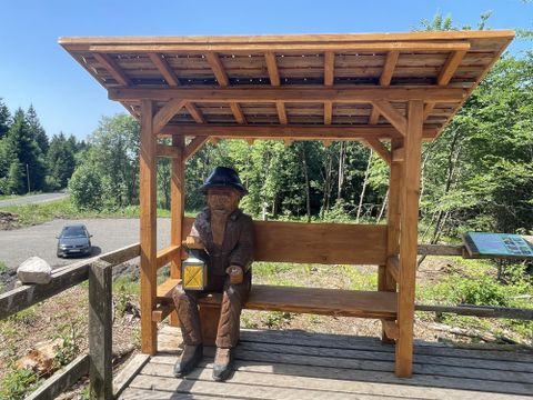 Un banc en bois avec une statue d'un homme tenant une lanterne. En arrière-plan, on peut voir des arbres et une rue.