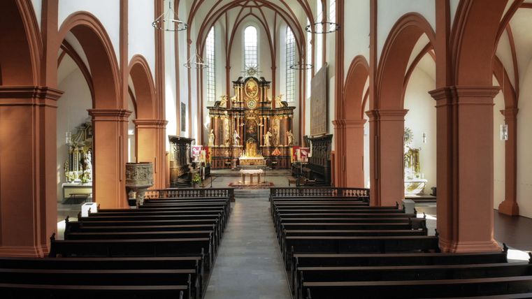 Eine Innenansicht einer Kirche mit hohen, gewölbten Decken und roten Bögen. Die Bänke sind in Reihen angeordnet und der Altar ist im Hintergrund sichtbar.