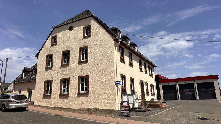 Ein dreistöckiges Gebäude mit einer hellen Fassade und mehreren Fenstern. Im Hintergrund ist eine Garage sichtbar.