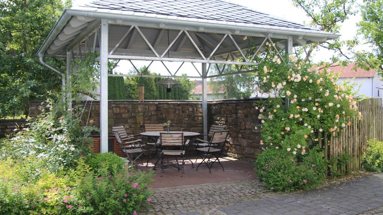 Eine gemütliche Pergola mit einem Tisch und Stühlen. Rundherum wachsen Blumen und Pflanzen.