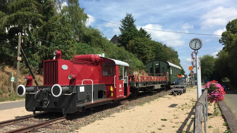 Eine rote Lokomotive steht an einem Gleis neben einem älteren Waggon. Im Hintergrund sind Bäume und ein Uhrturm zu sehen.