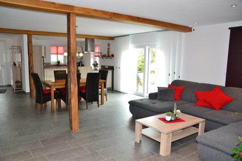 A modern living room with a cozy sofa and a wooden table. In the background, there is a dining area and large windows that let in plenty of light.