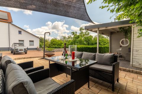 A cozy outdoor area with a black rattan sofa and a table. In the background, a well-kept garden and a bright, friendly atmosphere can be seen.