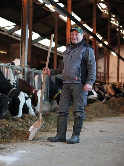 Ein Landwirt steht in einem Stall mit Kühen und hält eine Schaufel in der Hand. Er trägt Gummistiefel und Arbeitskleidung.