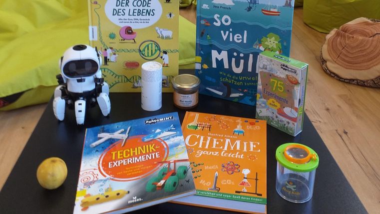 A table with various books, including titles on technology and chemistry. Additionally, there is a small robot, a glass, and an apple visible.