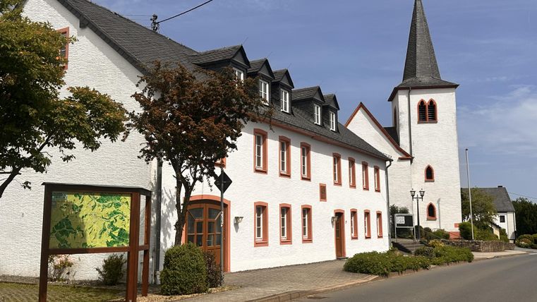 Ein charmantes Gebäude mit weißer Fassade und roten Akzenten steht neben einer Kirche mit spitzem Turm. Die Straße verläuft ruhig und ist von Bäumen und Sträuchern gesäumt.