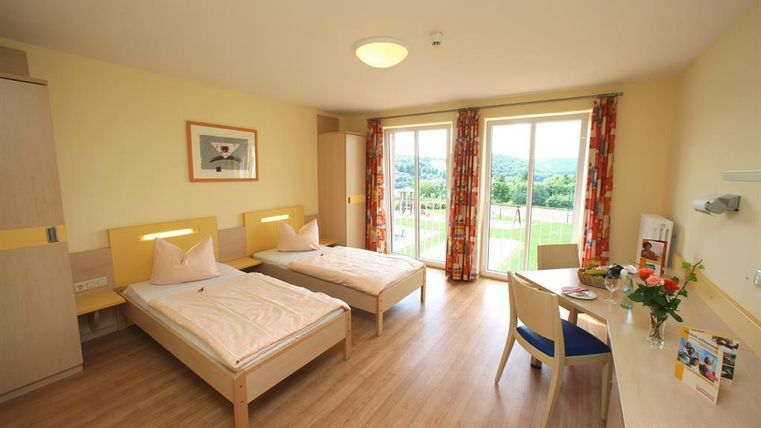 Ein helles Zimmer mit zwei Einzelbetten und großen Fenstern. Die Einrichtung ist modern und freundlich, mit einem kleinen Tisch und einer Vase mit Blumen.